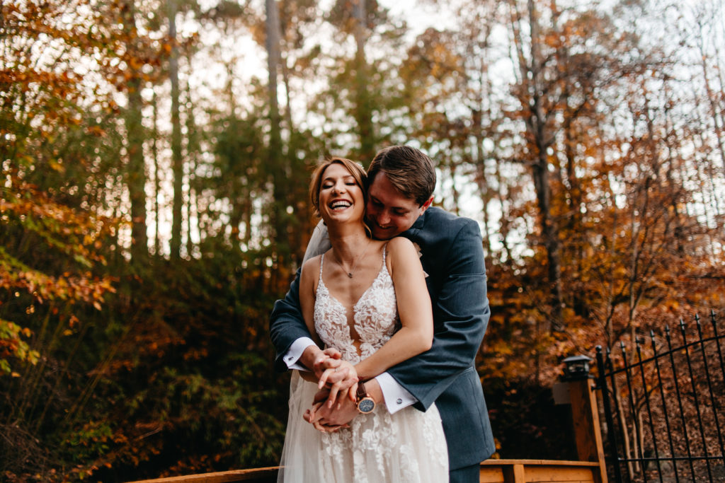 Groom holding bride close in laughter surrounded by fall trees 
